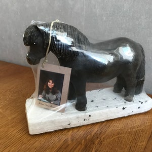 May include: A black horse figurine with a white base. The figurine is wrapped in plastic and has a tag with a picture of a woman and the text "Eichen Grove Farm".