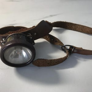 May include: Vintage brown leather miner's headlamp with a circular light and a brown leather strap. The headlamp has a dark brown, possibly bakelite, housing and a clear glass lens. The leather shows signs of wear, suggesting age and use.