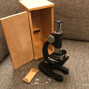 Könnte beinhalten: Ein schwarzes Mikroskop mit einem Holzkoffer. Das Mikroskop hat einen schwarzen Metallkörper und einen silbernen Knopf. Der Koffer ist aus hellem Holz und hat einen Klappdeckel.