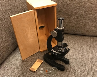Könnte beinhalten: Ein schwarzes Mikroskop mit einem Holzkoffer. Das Mikroskop hat einen schwarzen Metallkörper und einen silbernen Knopf. Der Koffer ist aus hellem Holz und hat einen Klappdeckel.