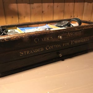 Puede incluir: Una caja de madera vintage para hilo de bordar, etiquetada como "Clark's Anchor Stranded Cotton for Embroidery". La caja tiene una tapa de cristal y cajones, con suministros de costura visibles en el interior.
