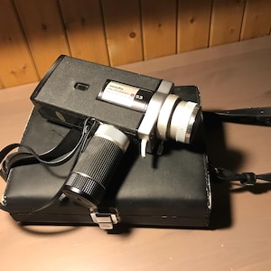 Può includere: Una cinepresa vintage Minolta Autopak 8 S3 Super 8 con corpo nero e obiettivo argentato. La fotocamera poggia su una custodia nera con un cinturino nero. Un obiettivo cilindrico è attaccato alla fotocamera.