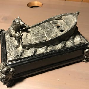 Op de afbeelding: Een decoratieve metalen asbak met een bootontwerp en een berenfiguur. De asbak heeft een zwarte basis met sierlijke hoekdetails en een zilverkleurige boot met drie cirkelvormige inkepingen.