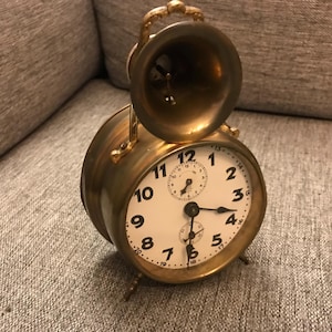 Op de afbeelding: Een vintage messing wekker met een witte wijzerplaat en zwarte cijfers. De klok heeft een bel bovenop en een kleine secondenwijzer. De klok staat op een grijze ondergrond.