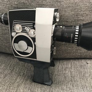 Op de afbeelding: Een vintage 8mm filmcamera in zwart en zilver met een zwarte lens en een zwarte lenskap. De camera heeft een zilveren body en een zwarte handgreep.