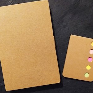 Peut inclure: Deux carnets en carton brun avec une rangée de cinq notes autocollantes colorées sur le plus petit carnet. Les notes autocollantes sont vertes, jaunes, roses, rose pâle et jaunes.
