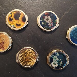 Peut inclure: Six miroirs de poche ronds à bordure dorée avec différents motifs floraux et naturels. Les motifs comprennent des tournesols, des fleurs, des notes de musique, des feuilles et des motifs floraux bleus.