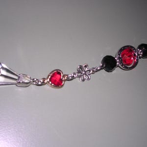 May include: A silver-toned necklace with a clear, faceted crystal pendant and a series of decorative elements. These include a red heart-shaped gem, a small flower, black beads, and a larger red gem in a silver setting. The necklace has a delicate chain.