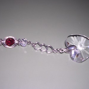 May include: A silver-toned heart-shaped crystal suncatcher with a faceted design. The suncatcher features a red, oval-shaped gem surrounded by clear, round beads and a delicate chain with heart-shaped links. The crystal heart is approximately 1.5 inches wide.