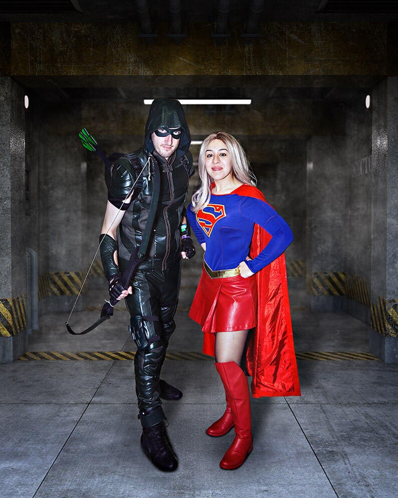 May include: A person dressed as Green Arrow stands next to a person dressed as Supergirl. Green Arrow is wearing a green hooded costume with a black vest and black pants. Supergirl is wearing a blue and red costume with a red cape. They are standing in a dark, industrial setting.