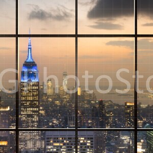 Puede incluir: Una vista de un horizonte urbano al atardecer a trav&eacute;s de una gran ventana con m&uacute;ltiples paneles. La ciudad est&aacute; iluminada con luces y el cielo es de un suave naranja y morado. El Empire State Building es visible en el centro de la imagen.