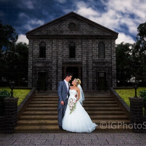 May include: A bride and groom stand on the steps of a stone building, smiling at each other. The building has a dark gray exterior and a large, arched doorway. The couple is dressed in traditional wedding attire, with the bride wearing a white gown and the groom wearing a dark suit. The sky is cloudy and overcast.