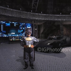 May include: A young person dressed as Batman stands in a dark, futuristic setting. They are wearing a black and gray costume with a yellow utility belt. A black car with a bat symbol on the hood is parked behind them. A large screen with blue and white graphics is in the background.