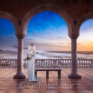 May include: A bride in a white wedding dress stands in a stone archway overlooking a beach with a blue sky and ocean in the background.
