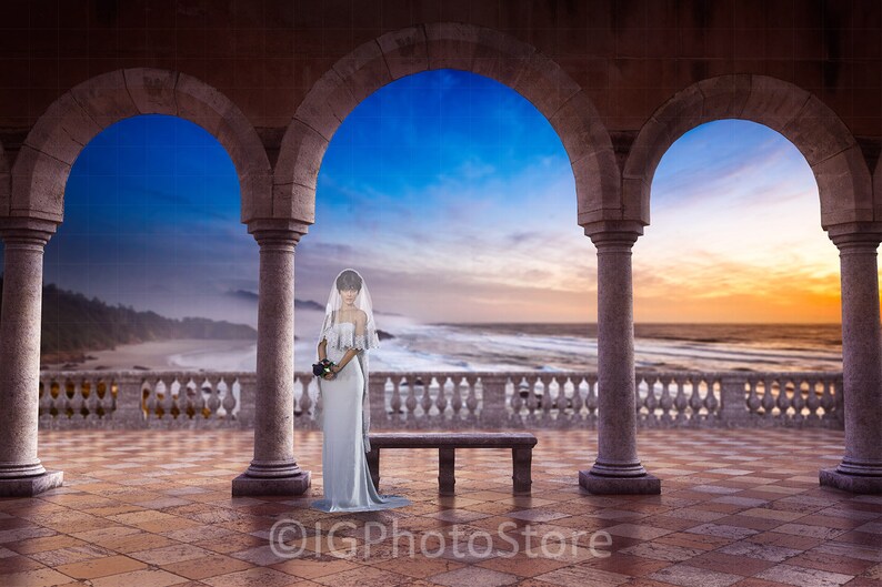 May include: A bride in a white wedding dress stands in a stone archway overlooking a beach with a blue sky and ocean in the background.