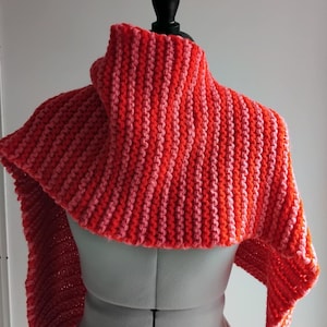 May include: A handmade, knitted shawl in a vibrant color palette of red and pink. The shawl is displayed on a mannequin, showcasing its ribbed texture and striped design. The warm tones suggest a cozy, winter accessory.
