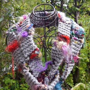 May include: A gray knitted scarf with colorful yarn accents and embellishments. The scarf is displayed on a wire mannequin.