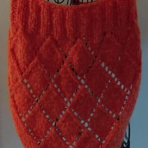 Peut inclure: Un châle tricoté orange avec un motif de losanges. Le châle est présenté sur un support en métal noir. Le tissu tricoté a un aspect texturé, et le motif de losanges présente des détails ajourés. La couleur est un orange vif et chaud.