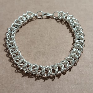 May include: A silver chainmail bracelet with a lobster clasp closure. The bracelet is made of small, interlocking rings, creating a textured, woven appearance. The bracelet is approximately 7 inches long.