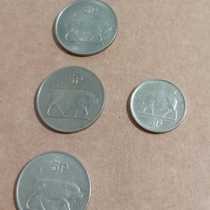 Puede incluir: Varias monedas plateadas de 5P están dispersas sobre una superficie beige. Cada moneda presenta la silueta de un toro y la inscripción '5P'. Las monedas son circulares y metálicas.