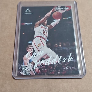 May include: A basketball trading card featuring Cam Reddish of the Atlanta Hawks in action, dunking the ball. The card is encased in a protective sleeve. The card has the Luminance logo and the text "RC" for Rookie Card.
