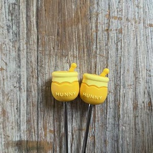 Puede incluir: Dos brochetas amarillas con forma de tarro de miel con la palabra "HUNNY" impresa. Las brochetas tienen una parte superior amarilla y un palito de metal plateado. El fondo es una superficie de madera.