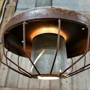 May include: A rustic, repurposed metal lampshade with a wire cage and a light bulb inside. The lampshade is made of rusty metal and has a weathered, industrial look.