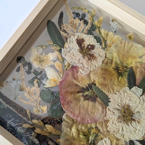 May include: A framed pressed flower arrangement featuring a selection of dried flowers in cream, beige and pink hues. The flowers are arranged in a bouquet style and are enclosed within a wooden frame. The inscription 'Neil & Ivana 15.09.2023' is visible in the bottom left corner of the frame.