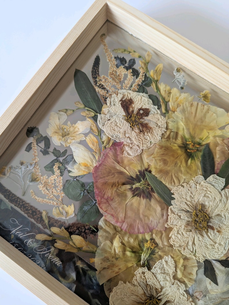 May include: A framed pressed flower arrangement featuring a selection of dried flowers in cream, beige and pink hues. The flowers are arranged in a bouquet style and are enclosed within a wooden frame. The inscription 'Neil & Ivana 15.09.2023' is visible in the bottom left corner of the frame.