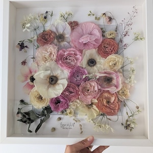 May include: A white frame containing a pressed flower arrangement. The flowers are mainly pink, white and peach. The text "Izzy & Will 18th May 2023" is written in black ink at the bottom of the frame.