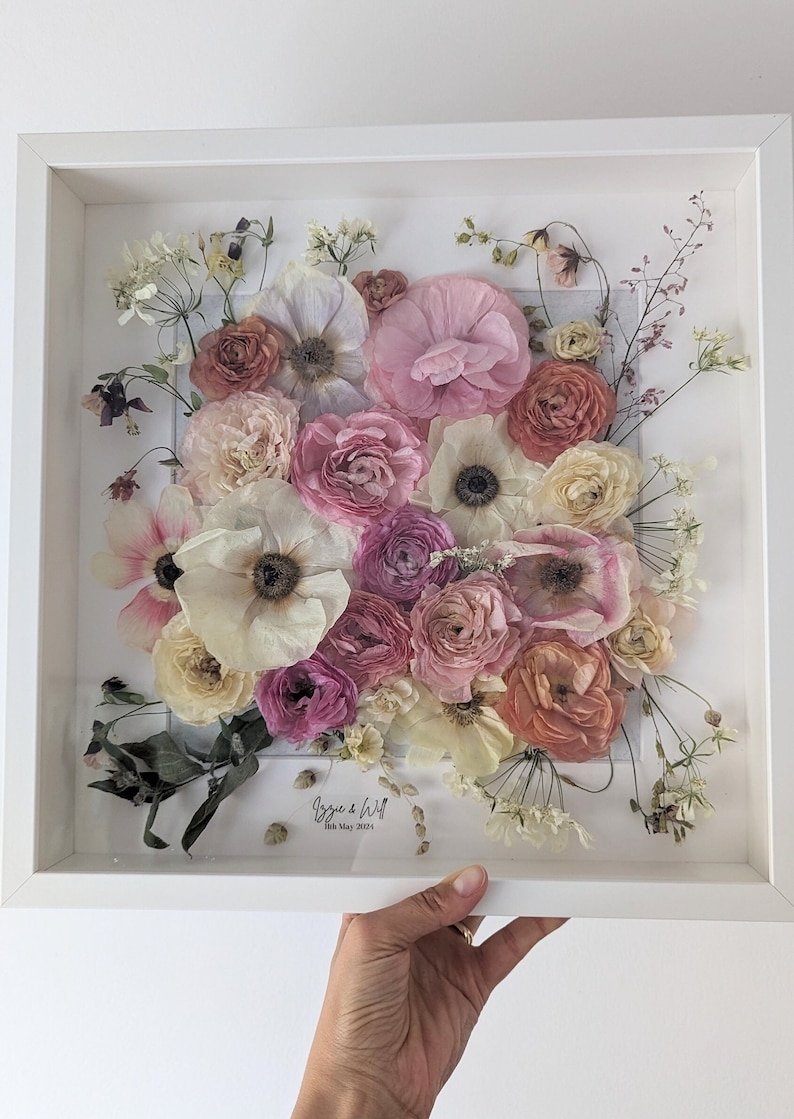May include: A white frame containing a pressed flower arrangement. The flowers are mainly pink, white and peach. The text "Izzy & Will 18th May 2023" is written in black ink at the bottom of the frame.