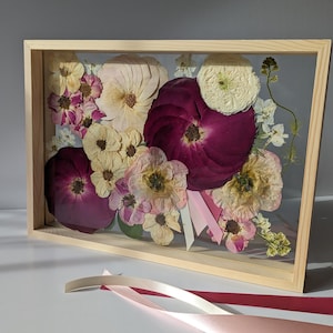 May include: A framed pressed flower arrangement featuring a selection of pink, white and burgundy flowers. The flowers are arranged in a bouquet shape and are pressed between two pieces of glass. The frame is made of wood and has a light brown finish.