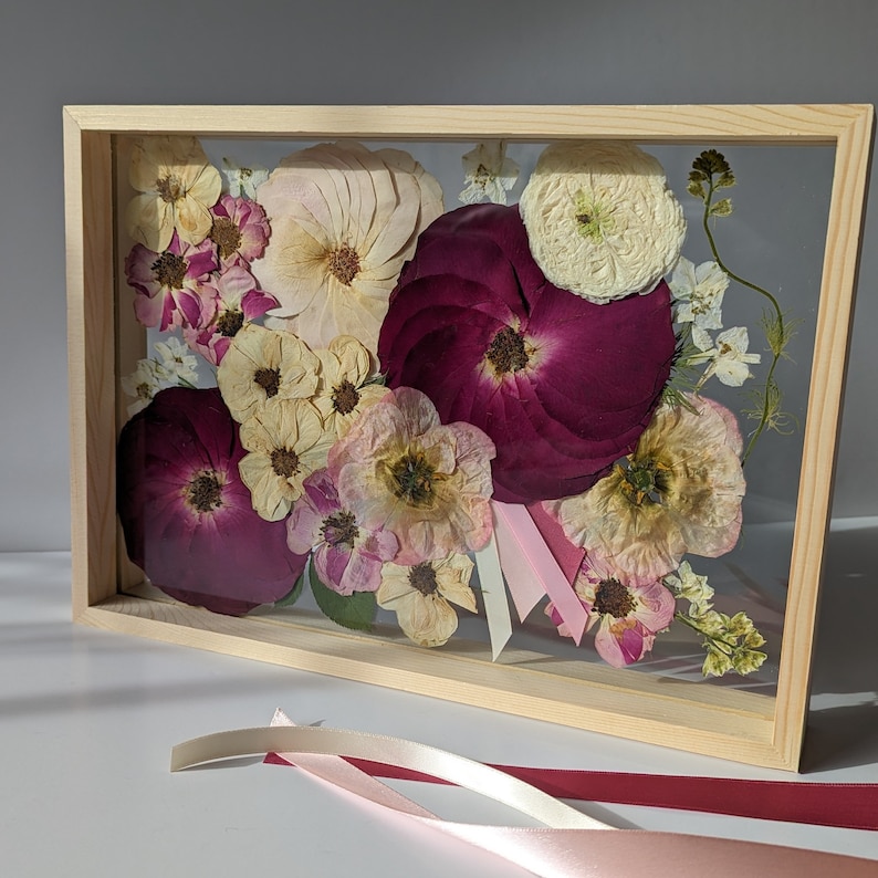 May include: A framed pressed flower arrangement featuring a selection of pink, white and burgundy flowers. The flowers are arranged in a bouquet shape and are pressed between two pieces of glass. The frame is made of wood and has a light brown finish.