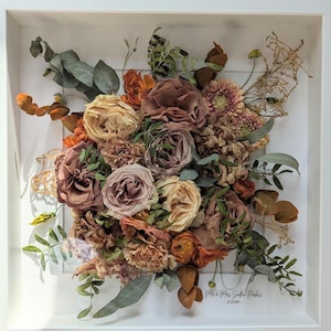 May include: A framed floral arrangement of dried flowers in peach, brown, and cream hues. The bouquet includes roses, carnations, and greenery, set against a white background within a white frame. The text "Mr. & Mrs. Smith Parker" and the date "12.27.2024" are visible.