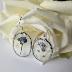 May include: A pair of silver earrings with a textured oval frame. Each earring features a pressed forget-me-not flower encased in clear resin.