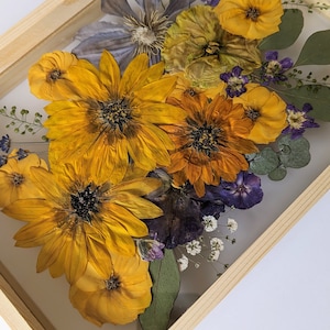 May include: A framed pressed flower arrangement featuring yellow, orange, and purple flowers. The flowers are arranged in a bouquet and are pressed between two pieces of glass. The frame is made of wood and has a light brown finish.