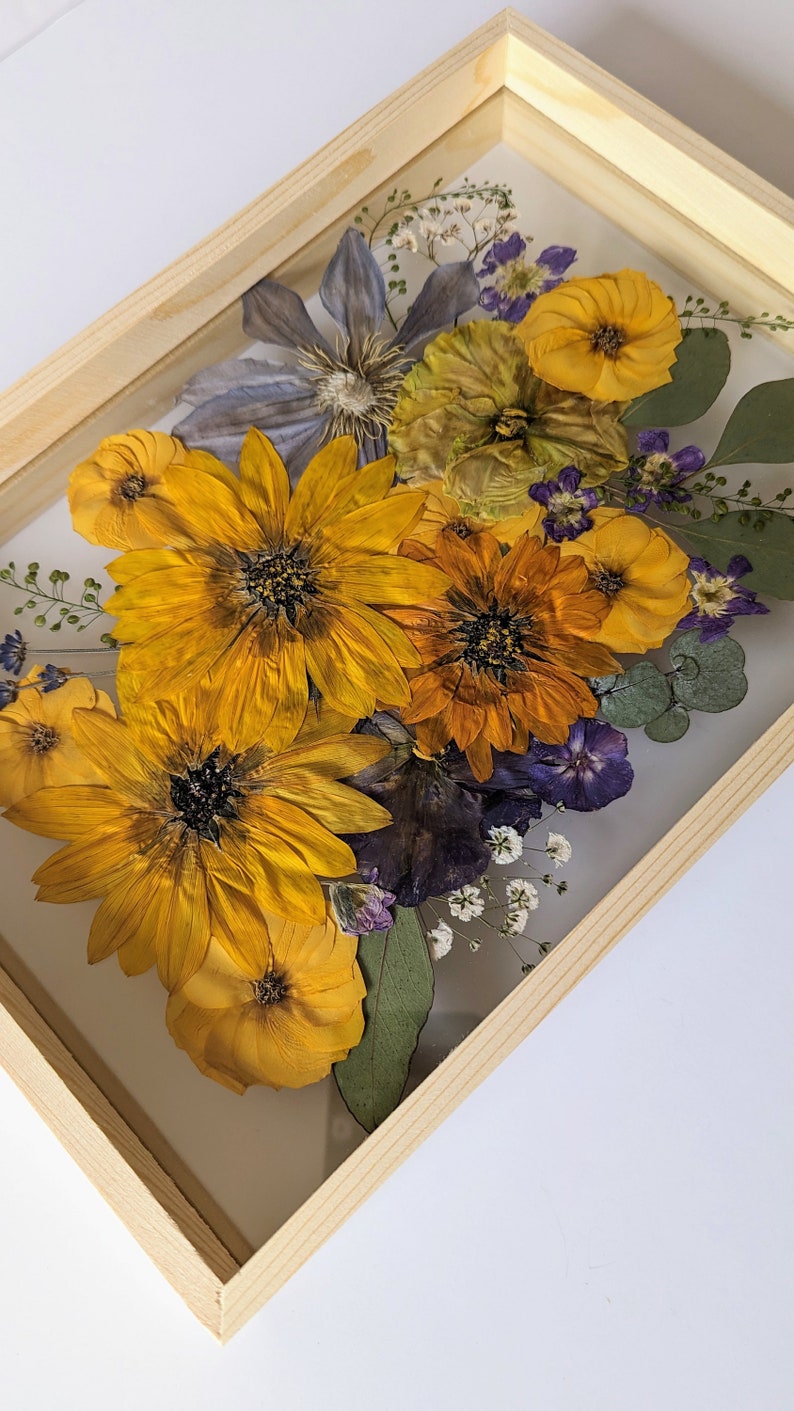 May include: A framed pressed flower arrangement featuring yellow, orange, and purple flowers. The flowers are arranged in a bouquet and are pressed between two pieces of glass. The frame is made of wood and has a light brown finish.