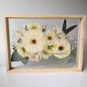 May include: A framed pressed flower arrangement featuring a selection of white and cream flowers, such as roses, lilies, and other delicate blooms. The flowers are arranged in a bouquet shape and are surrounded by green leaves and stems. The frame is made of natural wood and has a clear glass front.