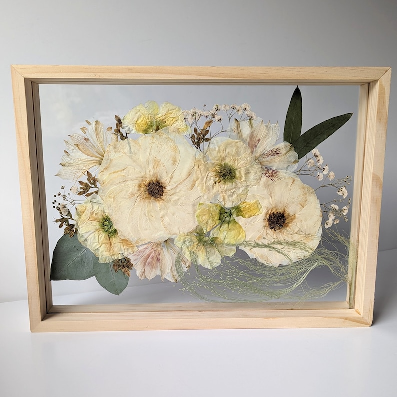 May include: A framed pressed flower arrangement featuring a selection of white and cream flowers, such as roses, lilies, and other delicate blooms. The flowers are arranged in a bouquet shape and are surrounded by green leaves and stems. The frame is made of natural wood and has a clear glass front.