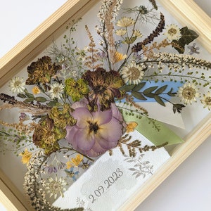 May include: A framed pressed flower arrangement with a light purple flower, dried grasses, and blue and green ribbons. The date "2.09.2023" is written on the white background.