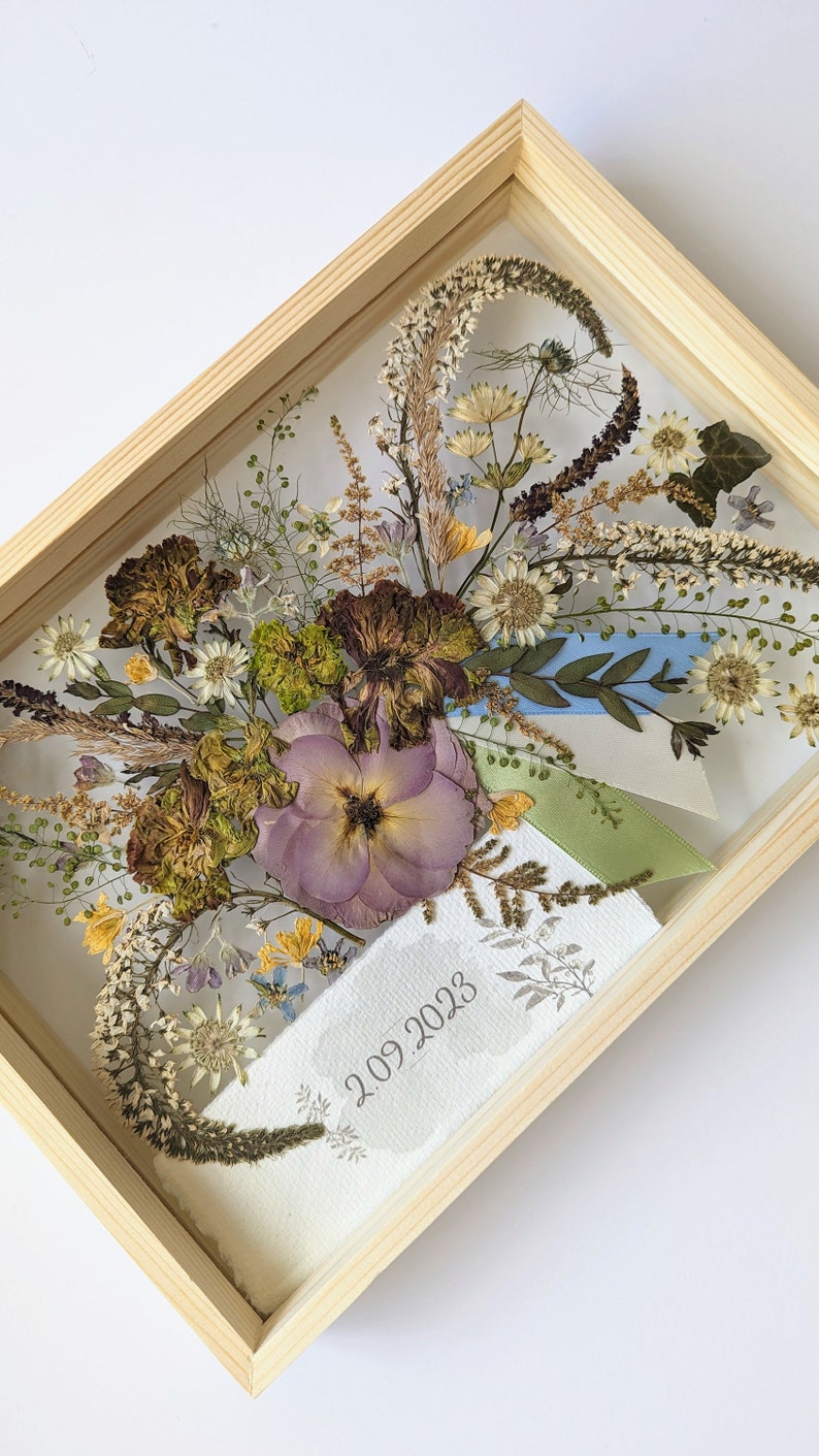 May include: A framed pressed flower arrangement with a light purple flower, dried grasses, and blue and green ribbons. The date "2.09.2023" is written on the white background.