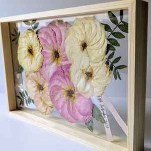 May include: A framed pressed flower arrangement with pink and cream flowers and green leaves. The date "11th September 2013" is written on a white ribbon.