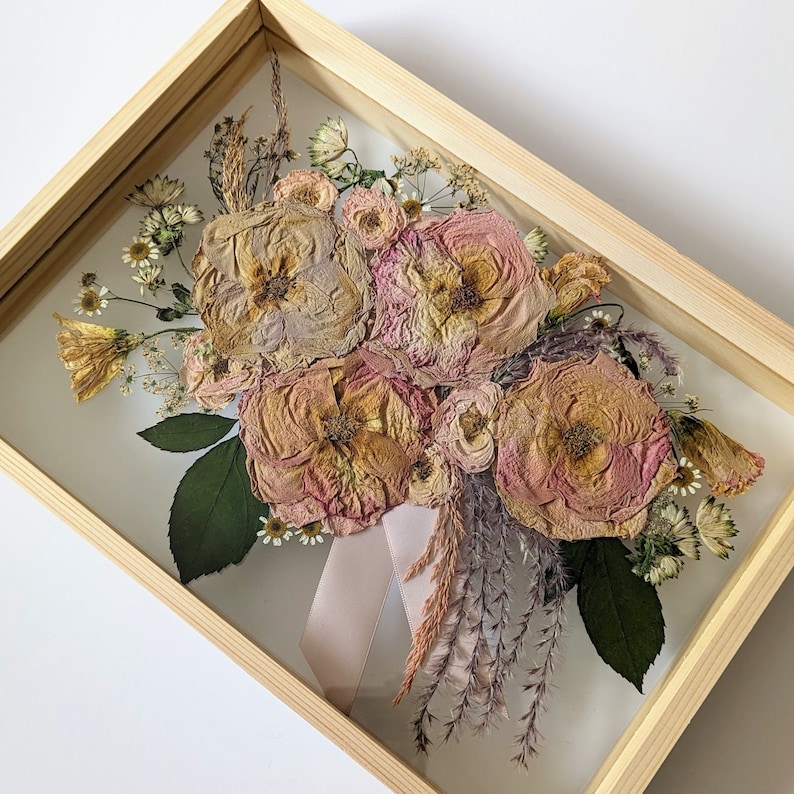 May include: A framed pressed flower bouquet featuring a selection of dried flowers, including roses, in shades of pink and beige. The bouquet is displayed in a wooden frame with a clear glass front.