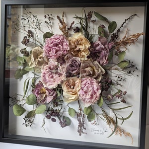 May include: A framed pressed flower arrangement featuring a mix of pink, white, and purple flowers, greenery, and dried grasses. The frame is black and has a white mount. The text "Eleanor & Luke 09.03.2024" is printed on the mount.