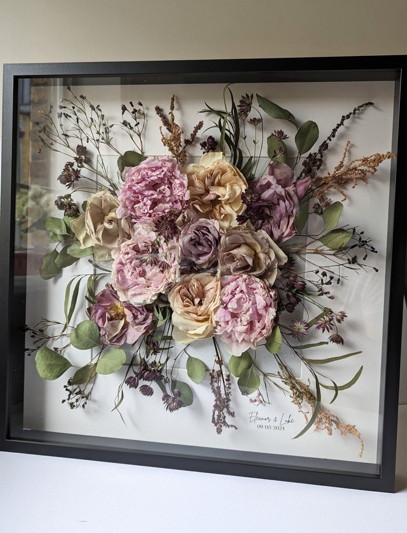 May include: A framed pressed flower arrangement featuring a mix of pink, white, and purple flowers, greenery, and dried grasses. The frame is black and has a white mount. The text "Eleanor & Luke 09.03.2024" is printed on the mount.