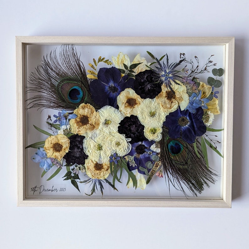May include: A framed pressed flower arrangement featuring white, blue and black flowers, green leaves and peacock feathers. The date "20th December 2023" is written in the bottom left corner.