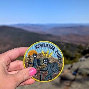 Może przedstawiać: Okrągła, żółto-niebieska naszywka haftowana z napisem "HUNGOVER HIKER" w żółtym kółku. Naszywka przedstawia dwie kreskówkowe postacie wędrujące z błyskawicami nad głową jednej z postaci.