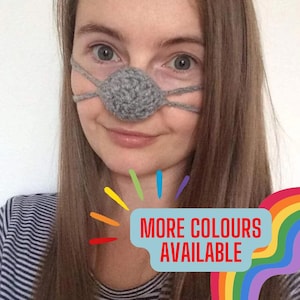 May include: A woman wearing a grey crocheted nose covering. The nose covering is a round ball with a strap that goes around the head. The text "MORE COLORS AVAILABLE" is in a speech bubble with a rainbow background.