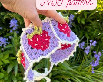 Pode incluir: Um gorro de crochê feito à mão com um design de morango. O gorro é feito com fio roxo, vermelho, verde e branco. O texto "PDF pattern" é exibido em um fundo rosa. O gorro é mostrado em frente a um fundo de folhagem verde e flores azuis.