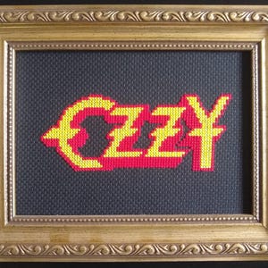 May include: Cross-stitched artwork featuring the name "OZZY" in yellow and red, set against a dark gray background. The framed piece is enclosed in a gold-colored, ornate frame, adding a decorative touch to the artwork.